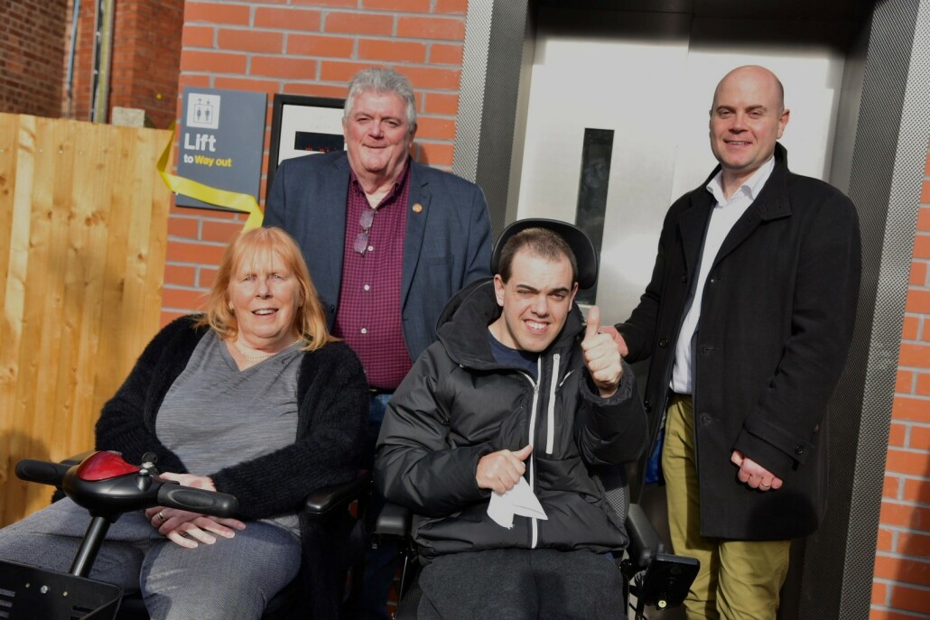 four people including two in wheelchairs smiling at the camera giving a thumbs up outside a lift 