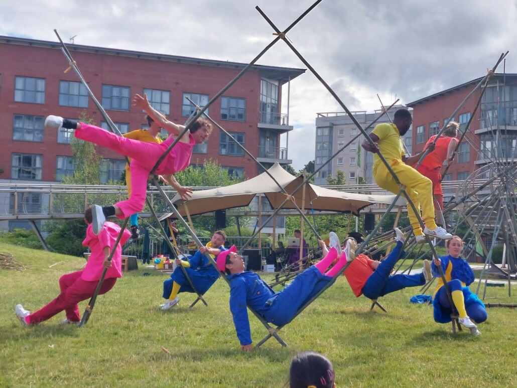 A group of performers dressed in different bright colours balance on a large structure made out of bamboo.