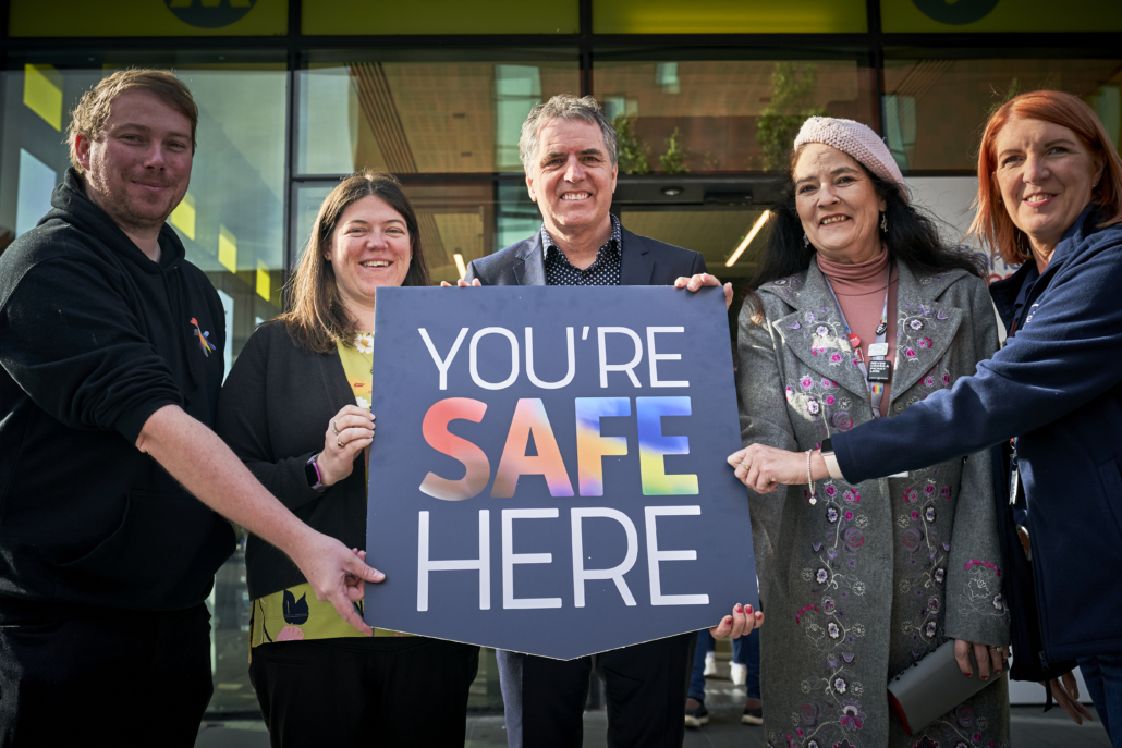 Andi Herring, Chief Executive Liverpool Pride Foundation; Emily Spurrell, Merseyside Police and Crime Commissioner; Liverpool City Region Mayor Steve Rotheram; Cllr Clare Carragher, Chair of the Travelsafe Board; Nikki Swanson, Travelsafe Officer all stand holding a 'you're safe here' sign.