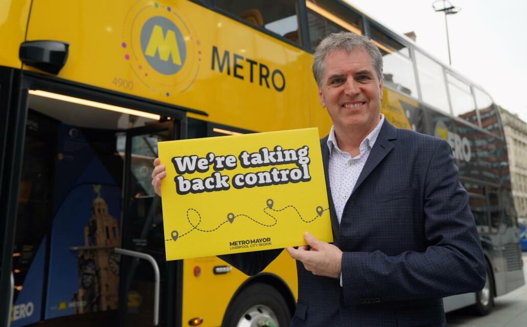 Mayor Steve Rotheram stands in front of a yellow bus holding a card saying 'we're taking back control'