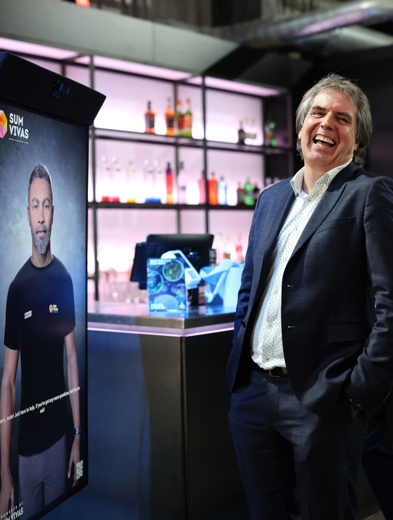 Metro Mayor Steve Rotheram looking at the camera and laughing and a screen in front of him displayed an AI avatar. The mayor is dressed in a grey suit with a white shirt.