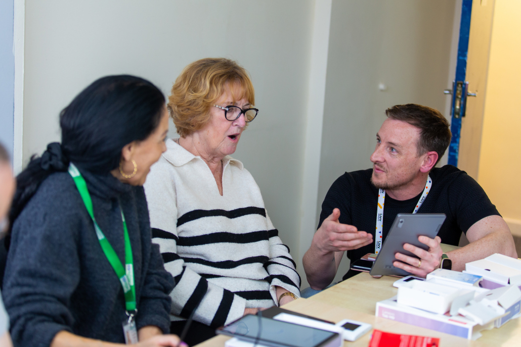 People sit in a class room style an a man using a tablet talks to two ladies to give them support