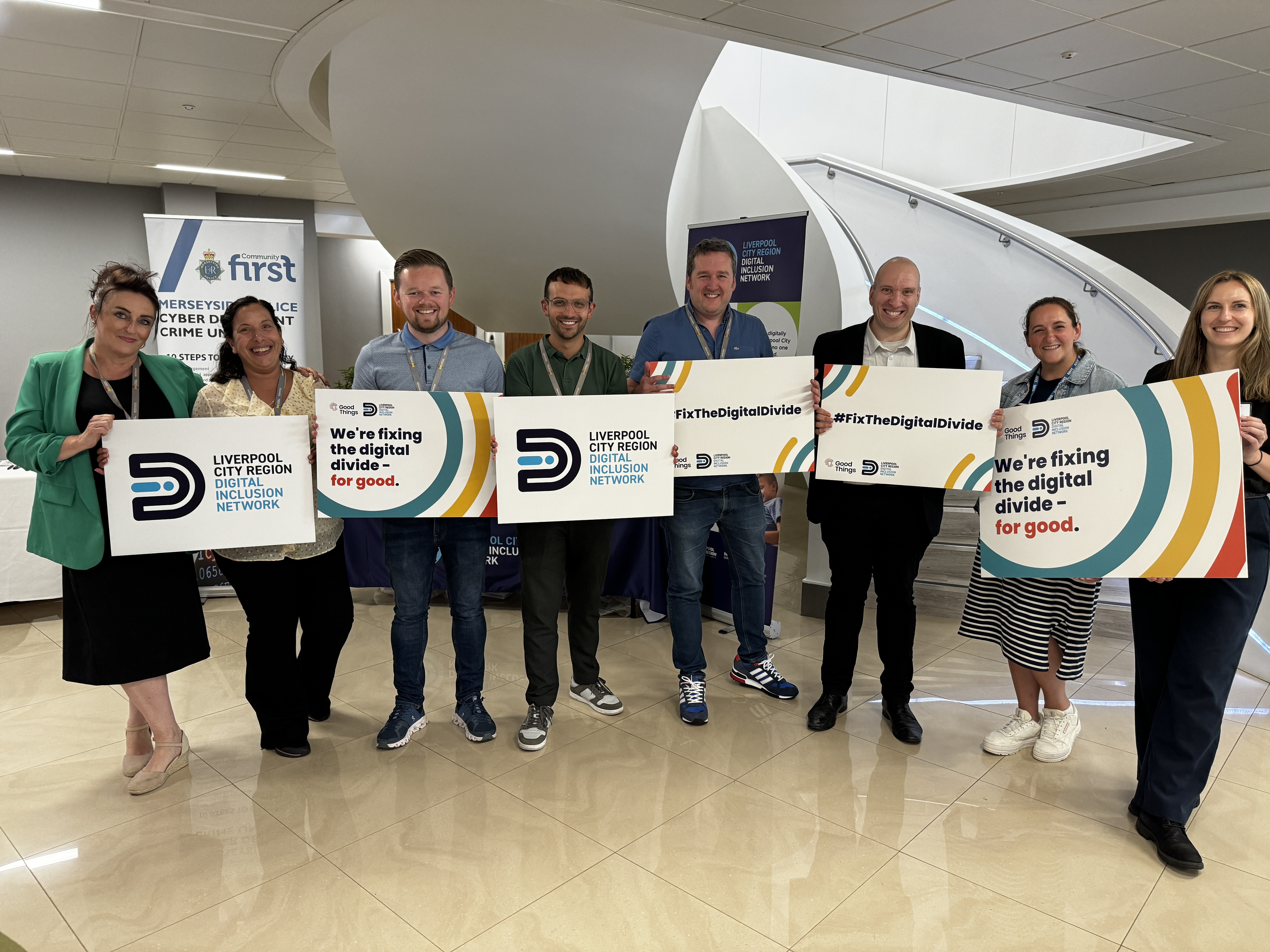 A group of 8 people stood smiling at the camera. They're each holding up a sign that all say different things on it such as 'We're fixing the digital divide', '#FixTheDigitalDivide' and the Digital Inclusion Network logo.