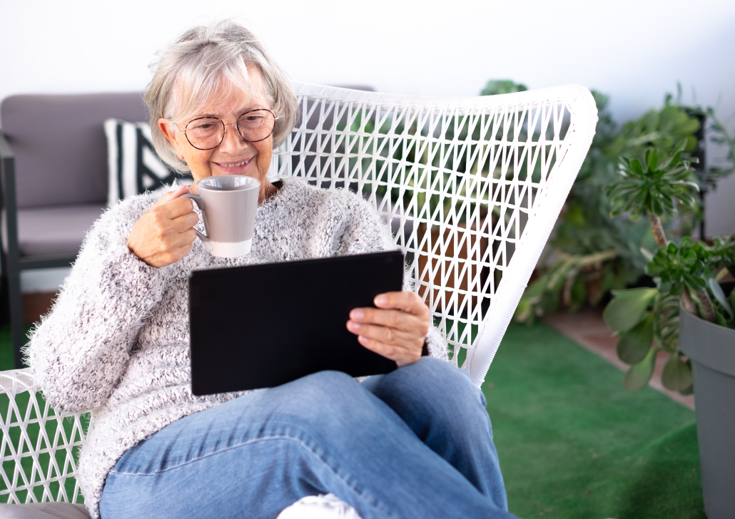 A woman who looks in her 60s sat looking at a digital tablet. The tablet is in one hand, a cup of tea is in the other. She is smiling.