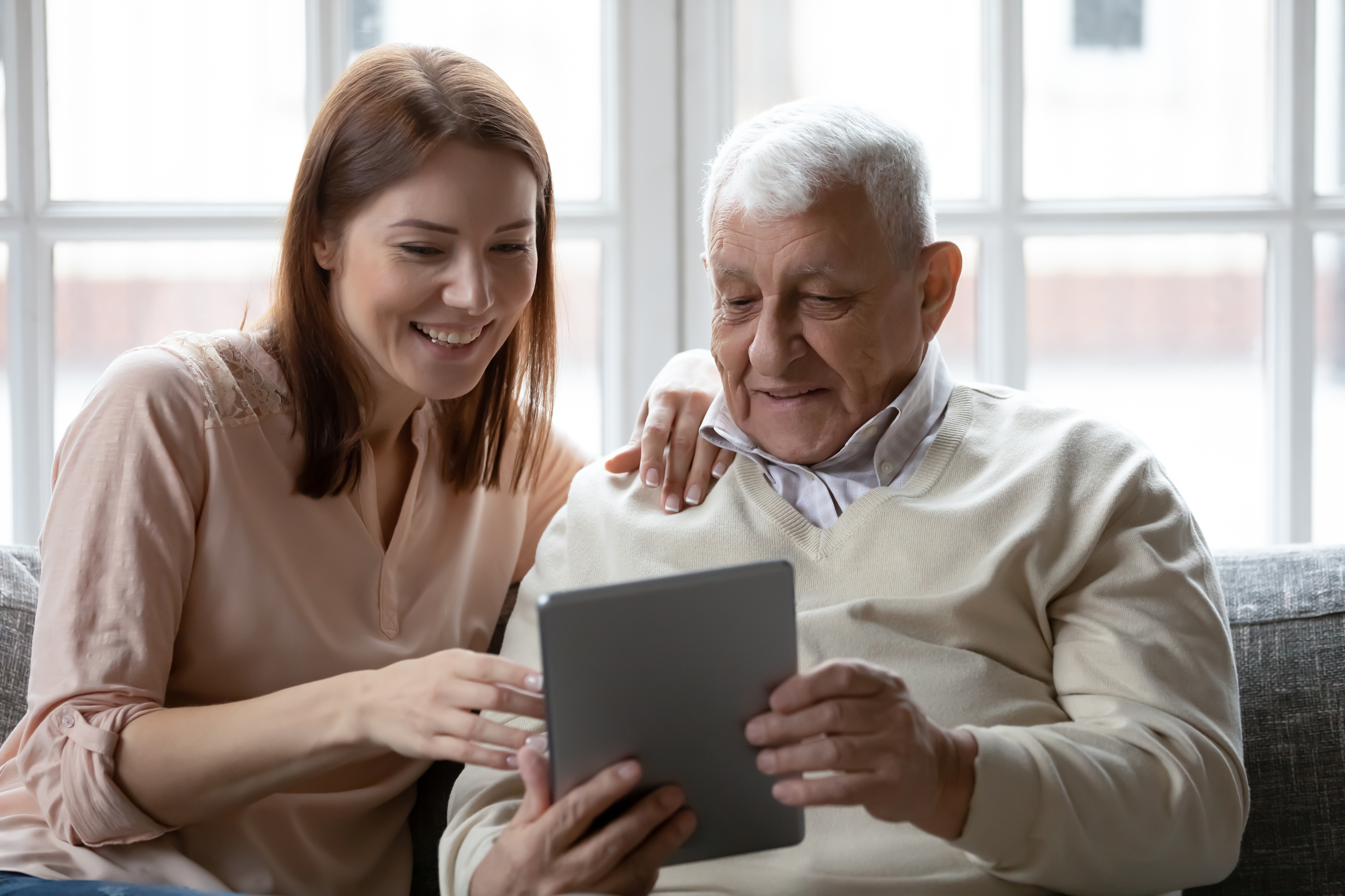 An old man holding a digital tablet with a young woman sat next to him. The woman is pointing at the tablet and appears to be supporting him.